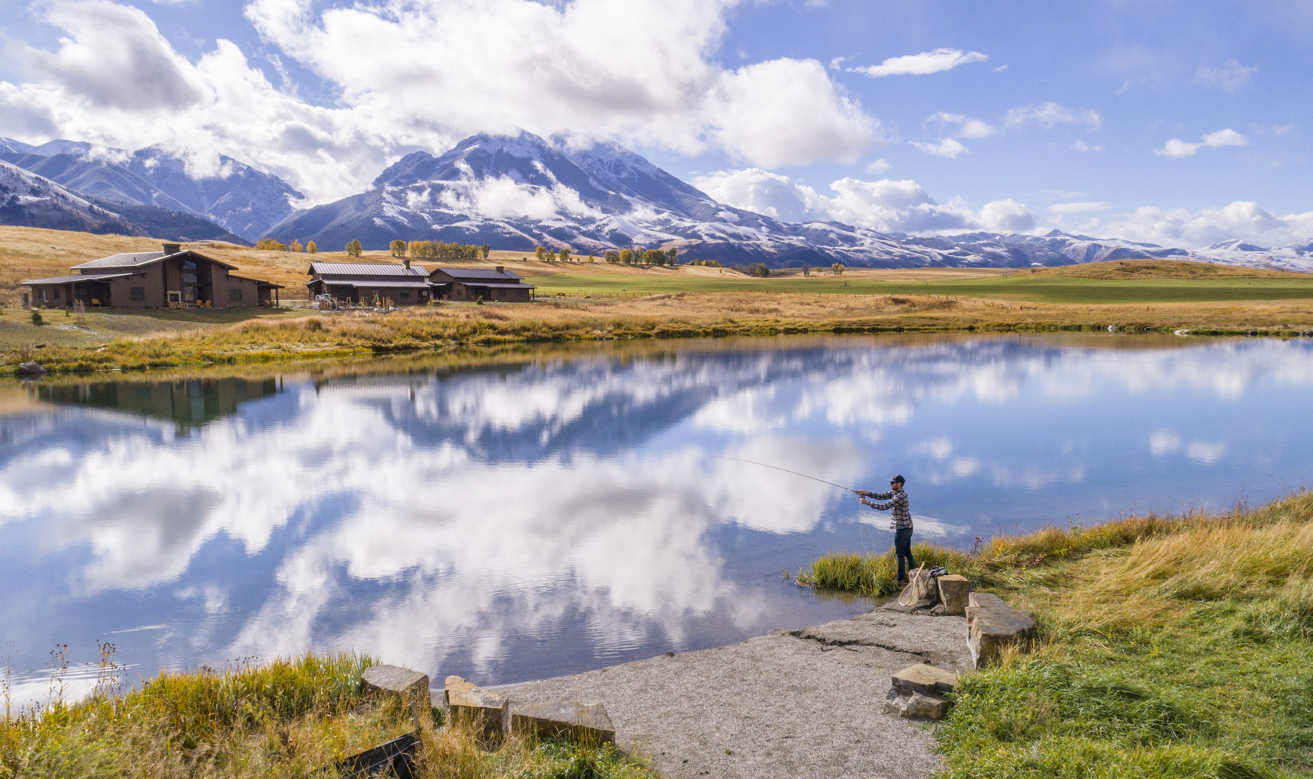 Person fishing in the pond with reflection of the lodge and sky in the water